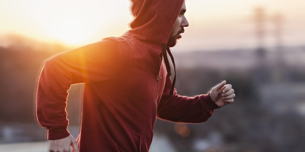 A picture of a man running and exercising beneath a clear sky, along a steal fence. He is wearing a red sweater with his hoodie pulled up and black shorts. In the distance below is some buildings and the sun is setting giving a nice warm light.
